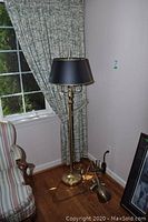 Tall brass floor lamp with black lampshade standing near window and curtain, on wood floor next to upholstered chair and artwork on floor.