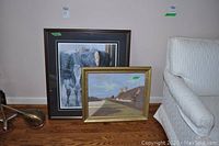 Two framed prints leaning against wall beside a white couch; larger winter scene print in dark frame with matting, smaller country scene print in gold frame showing rural house and road.