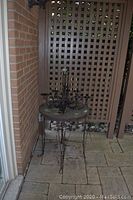 Metal table with round top and metal legs showing signs of wear and corrosion, with candle chandelier placed on top, set against brick wall and lattice background.