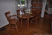 Full view of the wood oval dining table with four armless chairs and two armrest chairs around it, showing use marks and scratches on the floor and furniture surfaces.