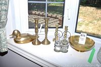 Overview showing brass duck loon tray, two brass candlesticks, corroded condiments holder, and brass bowl on window sill.