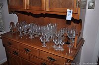 Photo showing an array of assorted glass and crystal glasses on a wooden dresser, including various styles of stemmed glasses.