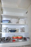 Kitchen cabinet shelves with assorted bowls, pitchers, plates, glass measuring cup, and serving dishes in various materials.