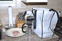 Photo of countertop showing electric kettle, French press, knife block, decorative plate, paper towel holder, and reusable shopping bag.
