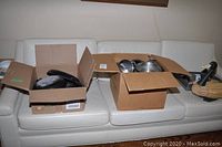 Boxes containing assorted used pots, pans, frying pans, and cookware on a white leather couch.