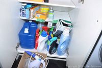 Overall view of white cabinet interior showing multiple cleaning supplies including large plastic jugs, spray bottles, boxed items, and a cardboard box with miscellaneous items on bottom shelf.