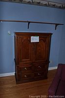 Front view of wooden armoire showing doors and four drawers with brass hardware, medium-brown wood finish, against blue wall.