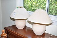 Two matching beige ceramic lamps with pleated cream fabric shades on a wooden dresser beneath a window.