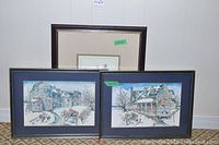 Three framed art prints arranged on floor against wall, two front prints show winter village scenes in Quebec with horse-drawn sleighs and snow-covered buildings, dark frames and blue mats, third print behind with lighter mat with stripes and dark wood frame.
