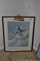 Full view of the framed print depicting a winter bird scene with a light fixture above the frame, leaning against wall on carpet.