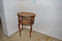 Two tiered wooden side table with oval surfaces, carved legs, and spindle accents, shown in corner against white panel wall and patterned floor.
