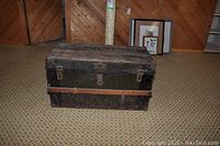 Antique wooden trunk with black paint and brown straps placed on patterned carpet against wooden panel wall.
