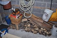 Wide view of stacked firewood on the floor with additional wood shavings and a plastic bucket nearby.