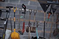 Long-handled garden tools including shovels, pitchfork, electric trimmer, and snow shovel against a concrete block wall
