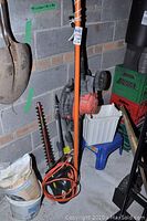 Photo showing orange pole hedge trimmer and other garden tools against cinder block wall