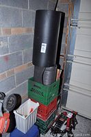 Three stacked plastic crates (green, red, white), with two rolled black mats on top and a small step stool visible inside the white crate. Items stored against a concrete wall in garage.