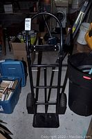 Black metal hand truck with curved handle and pneumatic tires shown upright in a storage area with various boxes around.