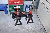 Photo showing Motomaster jack with attached handle and two jack stands with red ratchet handles, arranged on concrete floor near green crates and blue chair.