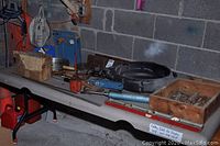 Vintage tools spread on a fold-out table including hammer, axe, saw blades, oil can, wooden boxes with parts, and a frying pan or metal tray on a grey fold table against a cinderblock basement wall