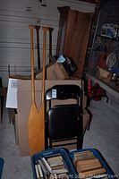 Photo showing two wooden paddles and two black folding chairs leaning against boxes in a garage.