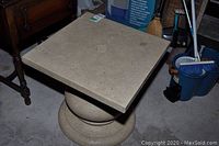 Photo showing square stone or concrete table with pedestal base, visible surface wear.