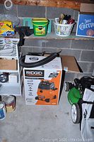 Wide shot of Ridgid shop vac box on floor near shelf and lawn mower