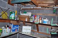 View of two wooden shelves with assorted garden and painting supplies including weed killers, insect repellents, spray paint, paint brushes, extension cords, and light bulbs. Shelves supported by wooden walls with items organized in baskets, boxes and loose distribution.