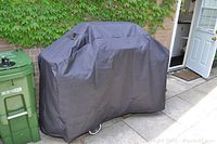 Broil King grill fully covered with new black cover, shown outside near a green trash bin and brick wall covered with ivy.