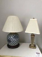 Two table lamps displayed side by side on a white surface: a blue and white Ming-style lamp with pleated beige shade and a gold painted lamp with lattice patterned base and cream shade.