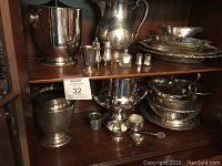 Wide view of multiple silver plated serving pieces grouped on a wooden shelf, showing pitchers, buckets, bowls, plates, small cups, and condiment containers.