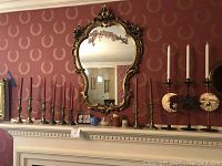 Large ornate gold-framed mirror mounted on wall above white mantel, showing damage and paint flaking. Display includes multiple brass candlesticks in pairs, assorted candles, and celestial-themed black metal candlestick holders with white taper candles.
