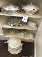 Photo of upper cabinet shelf with large and medium covered serving dishes, platters, plates, and butter dish displaying pink rose bud floral pattern and gold trim.