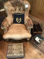 Front view of distressed leather chair with tufted back and nailhead trim, square needlepoint footstool in front, dark wooden magazine rack and decorative trash can to side on hardwood and oriental rug background.