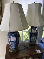 Both oriental style table lamps shown side by side on a wooden table near window, with newish off-white silk shades and bases featuring blue and pink floral design.