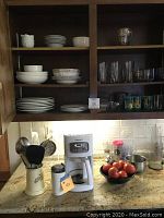 Kitchen cabinet open showing shelves with off white Sur La Table ceramic dishes including bowls and plates, assorted glassware including decorative and clear drinking glasses. Below on granite counter is white Cuisinart drip coffee maker and white coffee bean grinder, along with white crock holding kitchen utensils.
