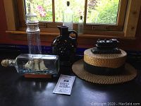 Photo showing a collection of antique and vintage curiosities on a dark surface in front of a window: tall glass bottle, square-shaped bottle with cork, dark jug, boater hat, and small model locomotive on hat