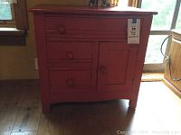 Front view of the antique red orange painted wooden chest showing three drawers on left and one cabinet door on right with round knobs, standing on wood floor in front of window.