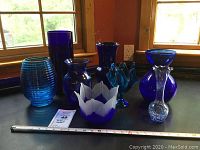 Eight assorted blue glass vases in varying styles and shades arranged on a black surface near a window, showcasing colors from cobalt to cerulean with mixed glass textures including ribbed and smooth finishes.