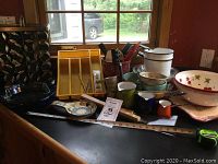 Longaberger crock, yellow spice rack, various Pyrex and Fire-King bakeware including bowls and pans, utensils including wooden spatula, ceramic and enamelware pitchers and bowls arranged on a countertop near a window.