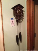 Full view of wooden cuckoo clock hanging on wall showing chalet-style roof, Roman numeral clock face, decorative carvings, and pinecone weights on chains.
