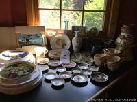 Photo showing a variety of china items on a table including plates, butter pats, teacups, and vases near a window.