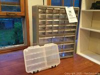 Photo showing large plastic storage rack with many small clear drawers stacked in rows and columns, behind a closed portable storage box lying flat on table.