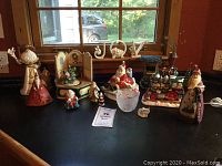 Wide shot of entire Christmas decor collection on black table by window showing multiple Santa figurines, JOY sign, Christmas tree calendar, candle holder, and Christmas train.