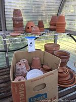 Photo showing multiple stacks of terracotta pots and several saucers on metal greenhouse shelf and inside cardboard box.