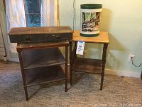 Two worn wooden side tables and a tall metal wastebasket with a wood duck illustration on its side shown on a carpeted floor with a wall in the background.