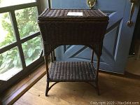 Front view of the dark brown vintage wicker stand with a woven top and lower shelf, placed near a window.