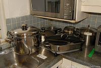 Wide view of kitchen counter showing multiple stainless steel pots and pans, IKEA skillet, and a Walmart electric grill pan with glass lid and temperature dial.