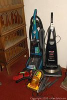 Photo showing four vacuum/floor cleaning units: Hoover Max Extract 60 steam cleaner, Panasonic EZ Bare Floor vacuum, yellow Eureka hand vac, and Dirt Devil hand vac, all placed on red carpet near wooden cabinet.