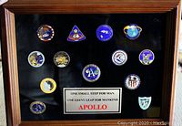 Full view of framed set of 13 Apollo program mission lapel pins arranged on black background with wooden frame and central plaque