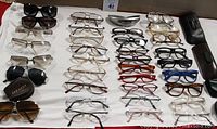 Overview of 34 pairs of assorted glasses and 4 eyeglass cases laid out on white cloth, including various frame styles, colors and sizes. Five eyeglass cases at right edge.